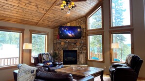 A spacious living room with a stone fireplace, leather sofas, a large TV, and large windows offering views of the outdoors. - Hot Tub, Mtn Views & Starlink WiFi - Wildlife on Site, Pet-Friendly (Fairplay)