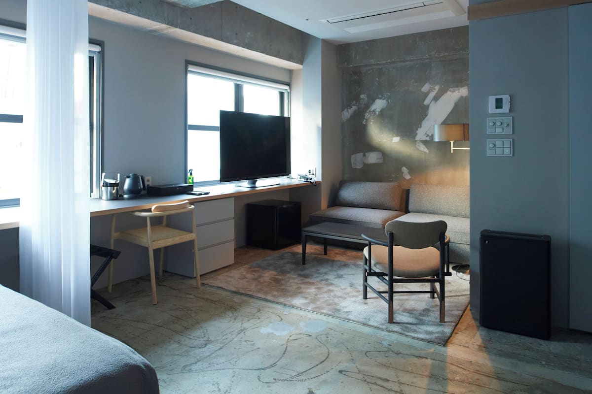 A modern hotel room with a large window, a flat-screen TV, a desk with a chair, a sofa, and a small refrigerator.