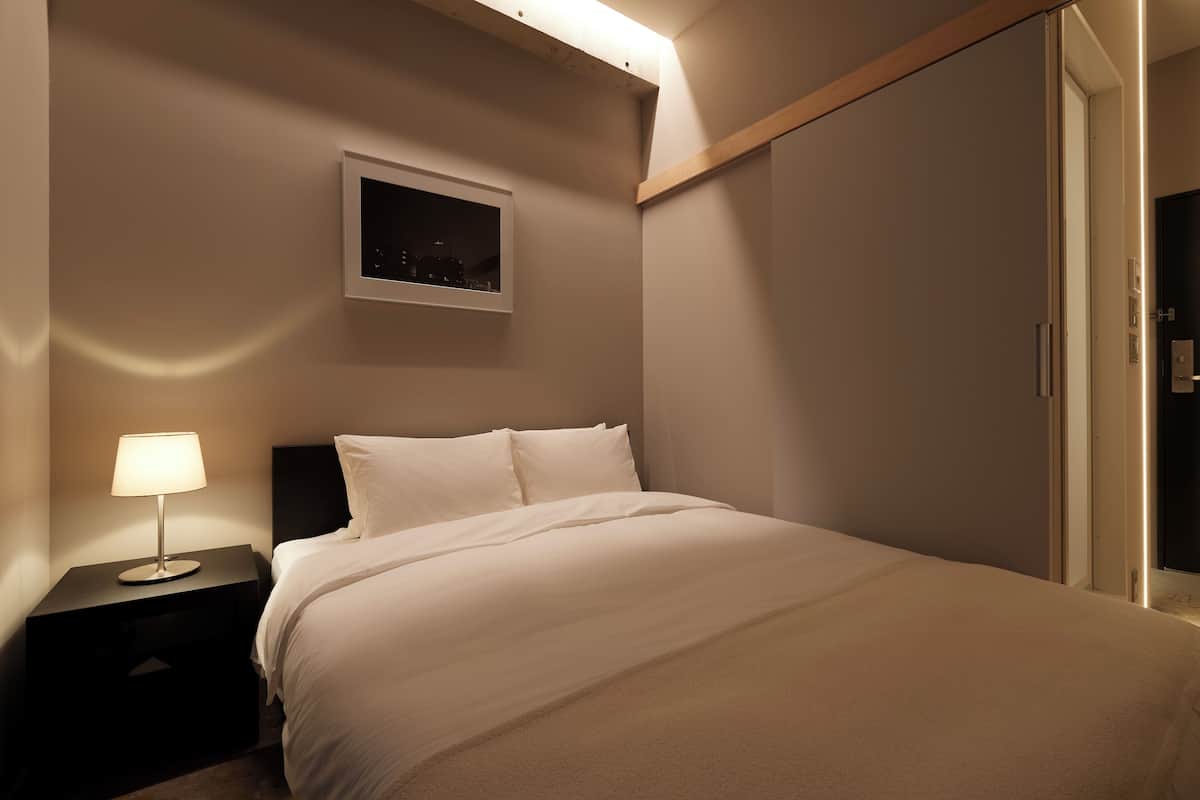 A neatly made bed in a hotel room with a nightstand, a wall-mounted picture, and a door with a light strip.