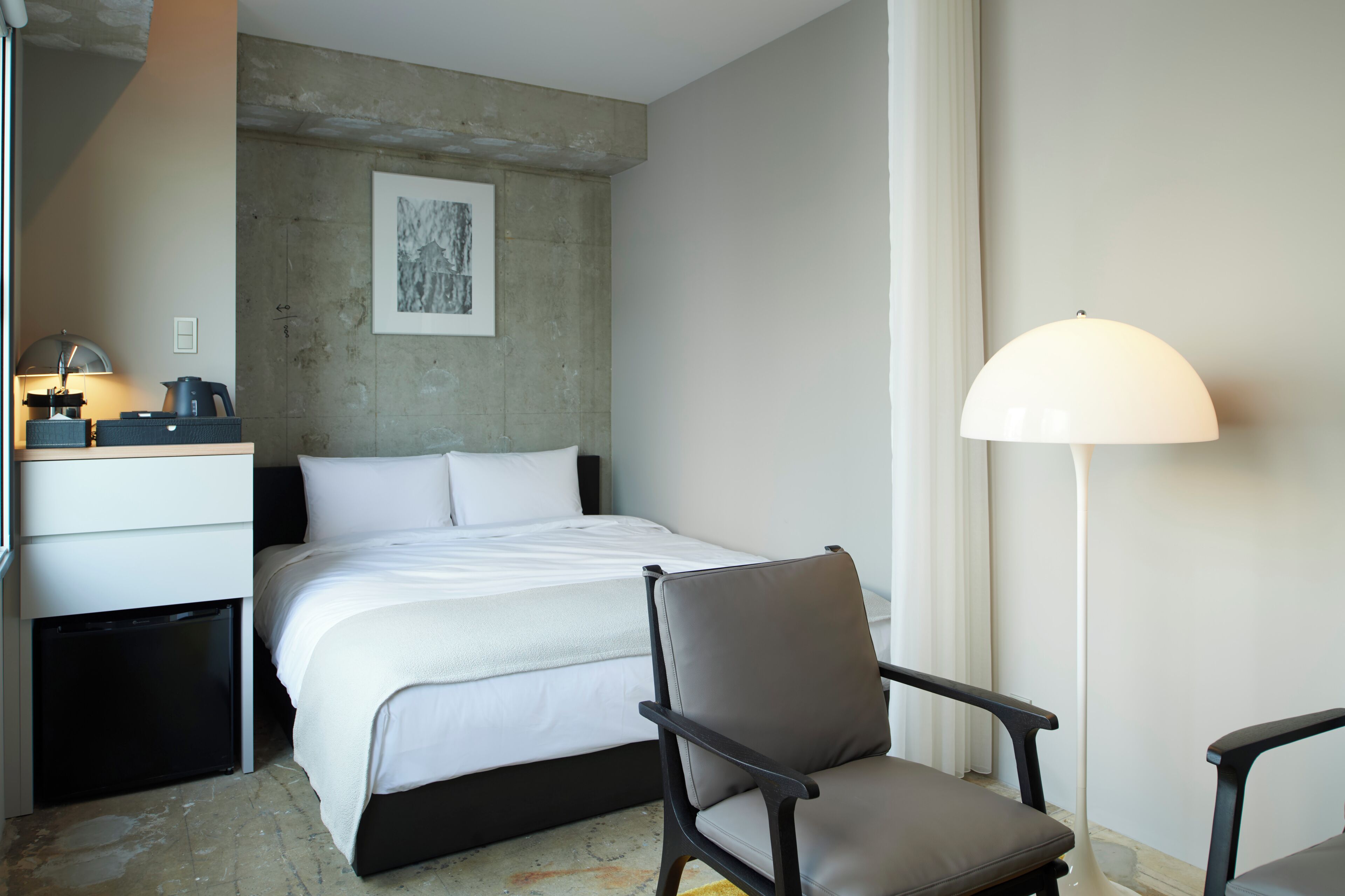 Et hotelværelse med en seng, en stol, et skrivebord med lampe og et billede på væggen.