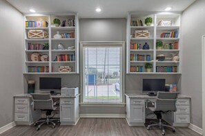 A study room with two desks, each with a computer and chair, flanked by bookshelves filled with books and decorative items.