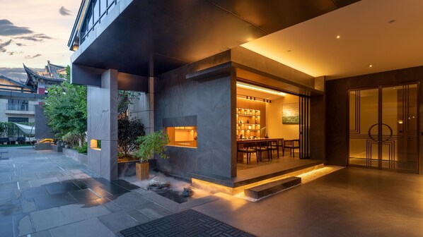 Un bâtiment moderne avec une entrée en verre, un espace repas avec des tables et des chaises, et une petite fontaine en premier plan.