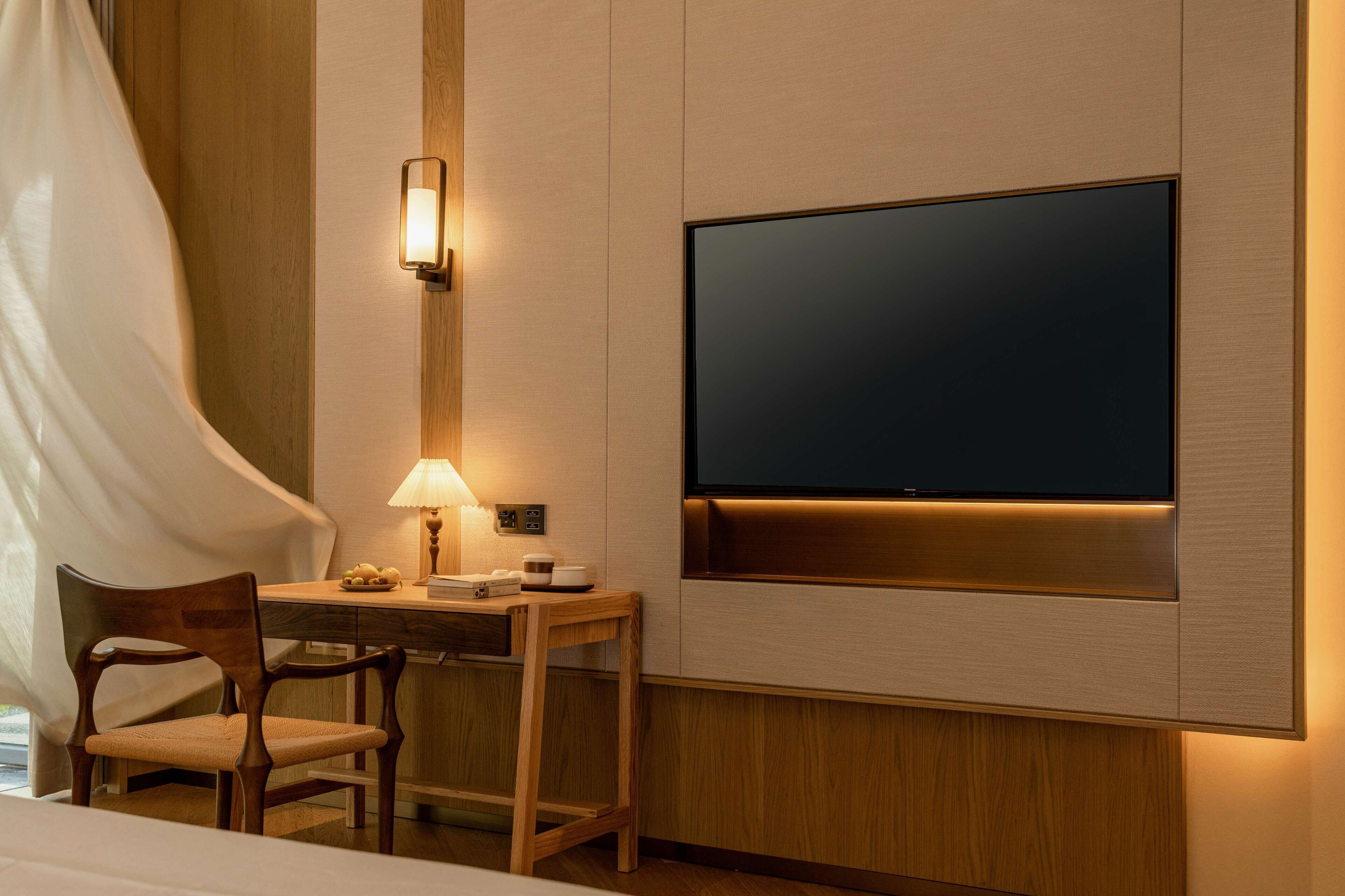 Habitación de hotel con cama, escritorio con lámpara, televisor montado en la pared y una silla.