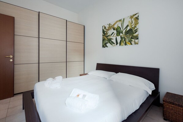 A neatly made bed with white linens and towels, a wooden wardrobe, and a wall-mounted plant.