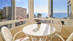 Un balcon avec une table ronde et des chaises en osier, donnant sur une plage et des immeubles.