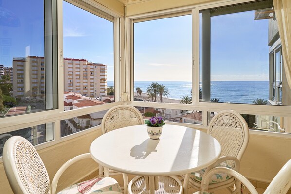 Un balcón con una mesa redonda y sillas de mimbre, con vista a una playa y edificios.