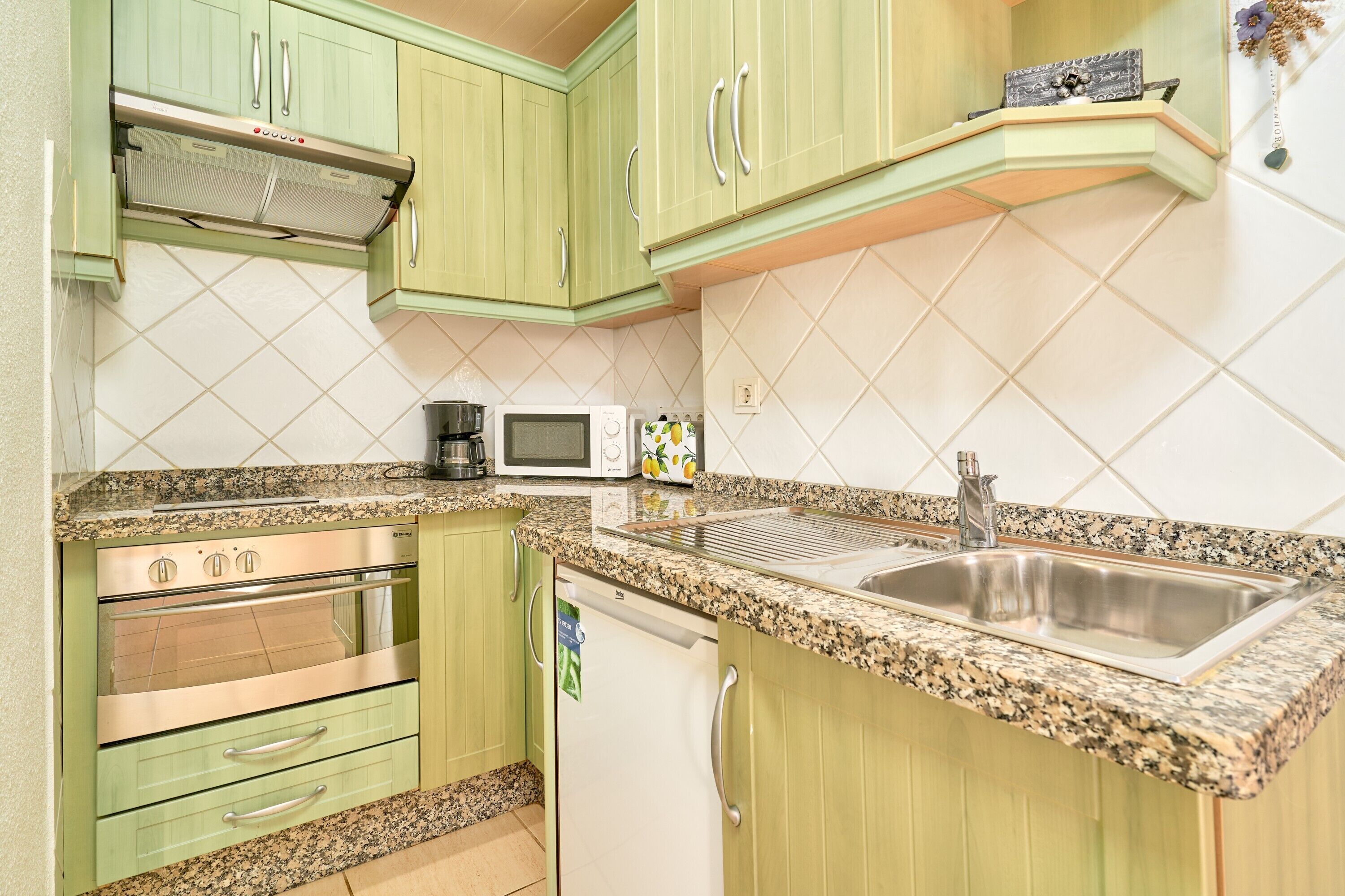 Una cocina con gabinetes verdes, un lavaplatos de acero inoxidable, un microondas y un lavavajillas.