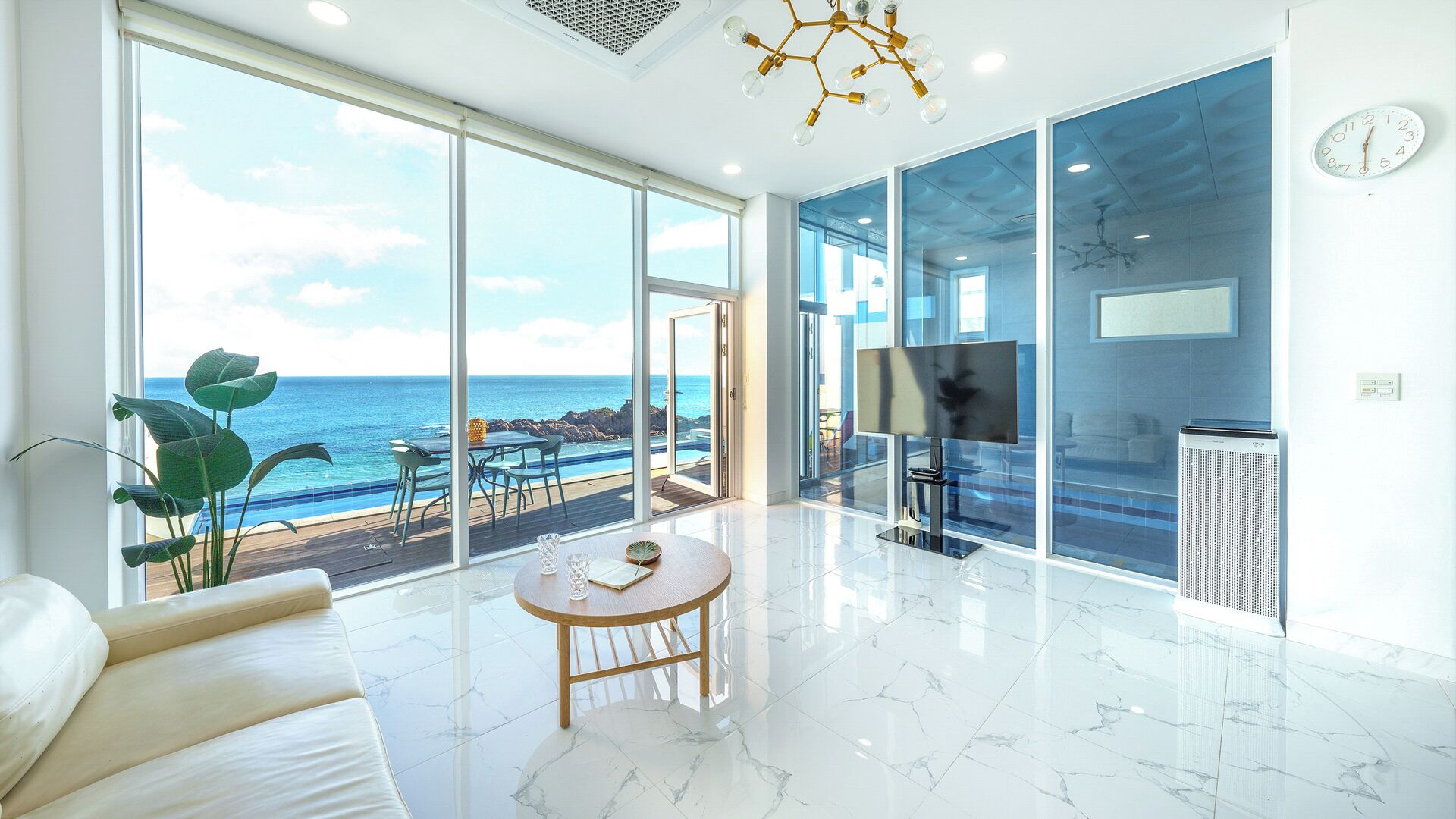 Ein modernes Wohnzimmer mit einem großen Glastür, der auf einen Balkon mit Meerblick führt, einem Flachbildfernseher und einem Marmorboden.