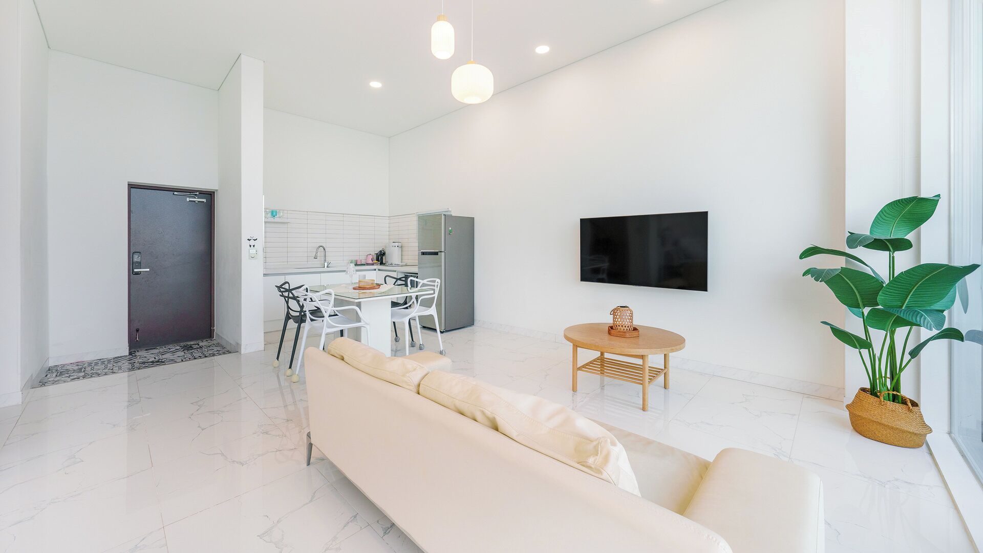 Una sala de estar moderna con un sofá, un área de comedor con sillas, una cocina al fondo, un televisor de pantalla plana montado en la pared y una planta en maceta.