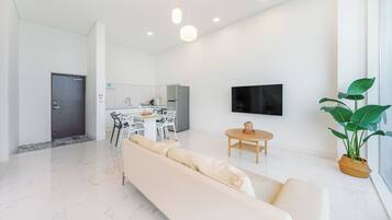 Una sala de estar moderna con un sofá, un área de comedor con sillas, una cocina al fondo, un televisor de pantalla plana montado en la pared y una planta en maceta.