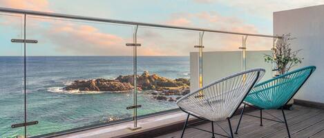 A balcony with a view of the ocean and rocky outcrops, featuring two chairs and a glass railing.