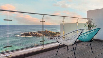 A balcony with a view of the ocean and rocky outcrops, featuring two chairs and a glass railing.
