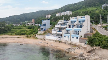 A coastal building with multiple balconies, a sandy beach, and a forested hillside in the background.