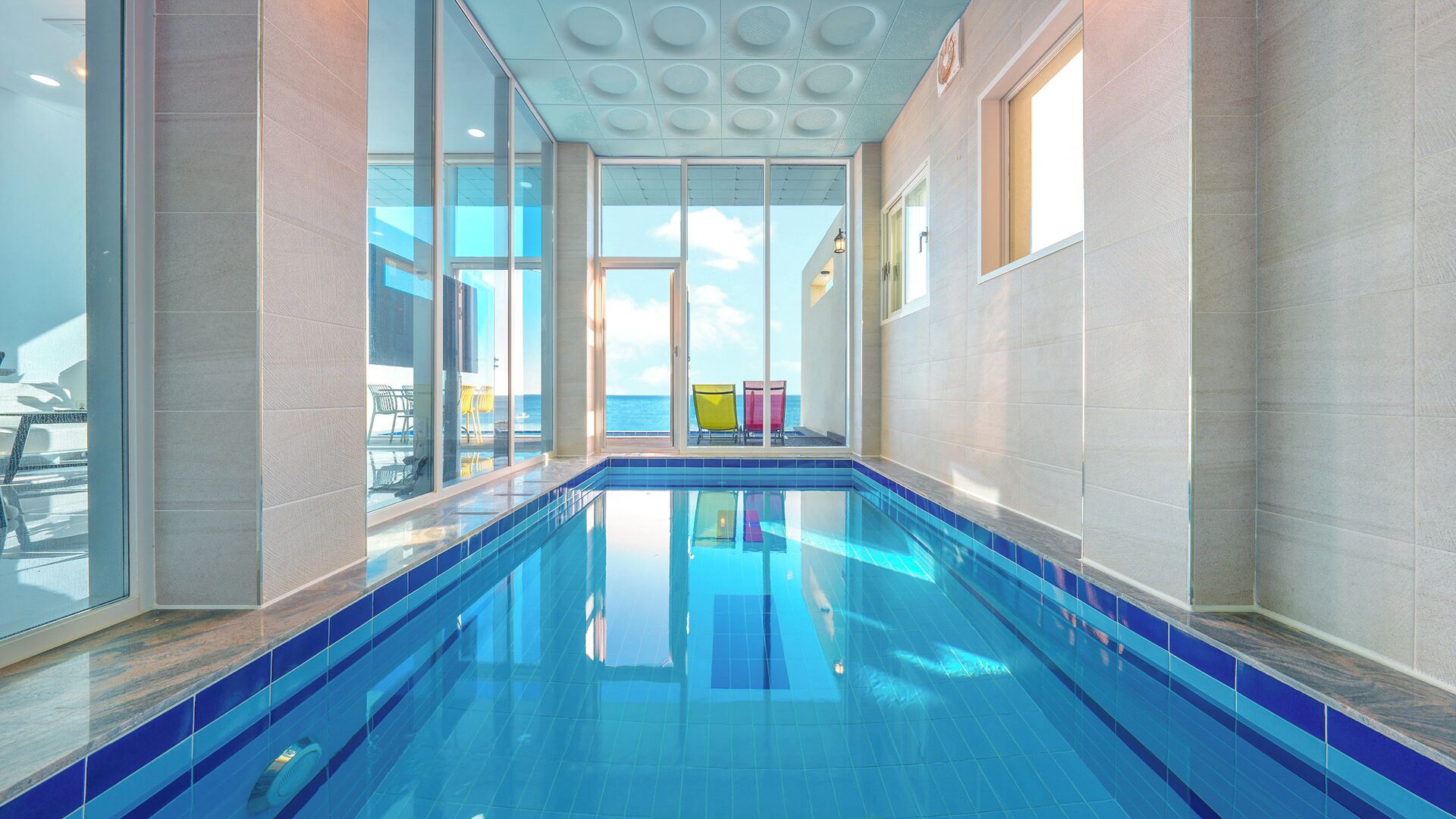 An indoor swimming pool with clear glass walls, a tiled floor, and a view of the sea through the windows.
