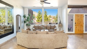 A spacious living room with a large sofa, a ceiling fan, and large windows offering a view of trees and mountains.