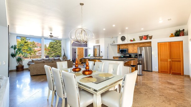 A spacious living area with a dining table, white chairs, and a chandelier. There is a kitchen in the background with wooden cabinets and a refrigerator.