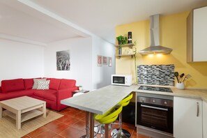 A compact kitchen with a microwave, stove, and oven, a red sofa, a small table, and a tiled backsplash.