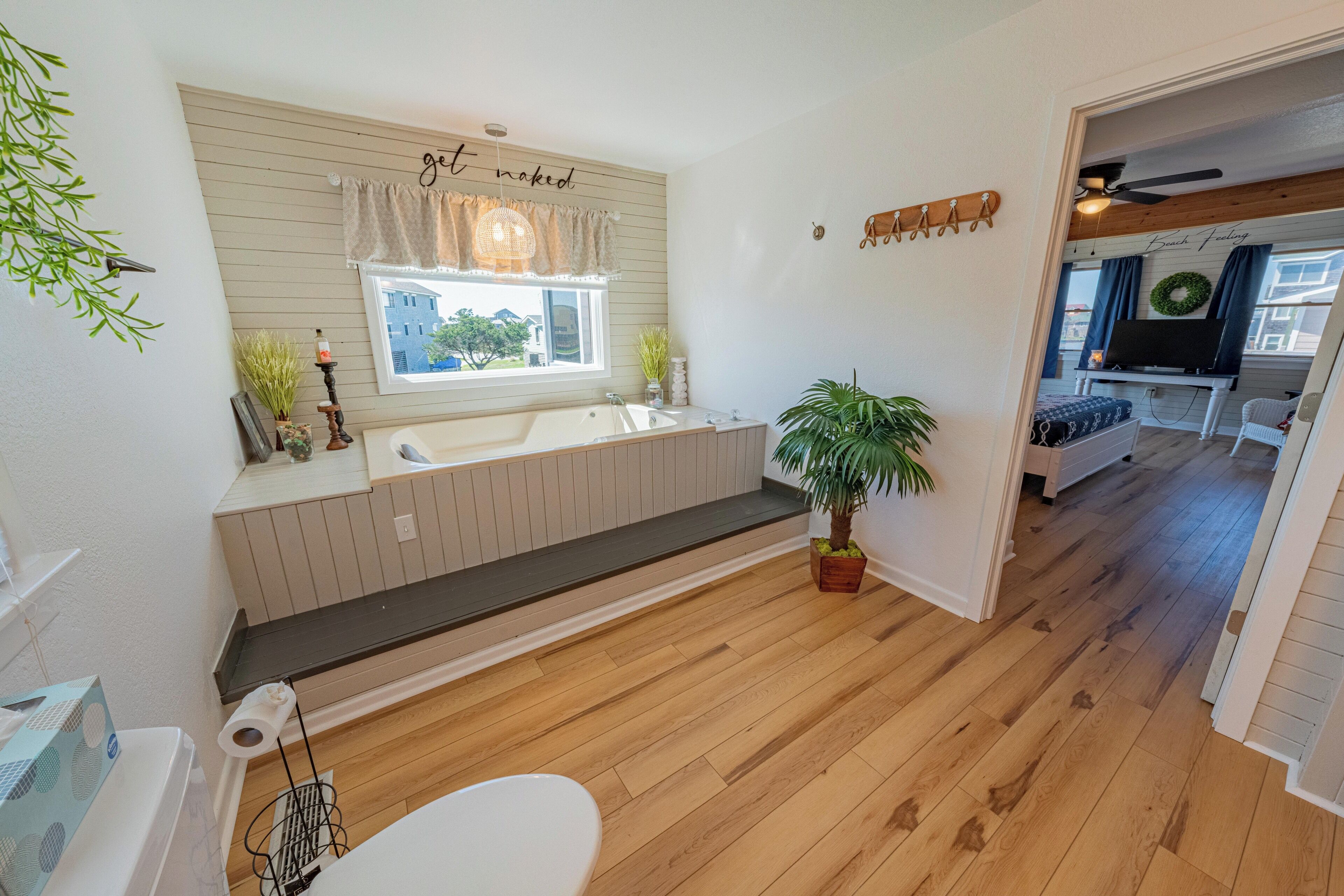 A modern bathroom with a bathtub, sink, and toilet. There is a window with a view of the outside, a ceiling fan, and a plant.