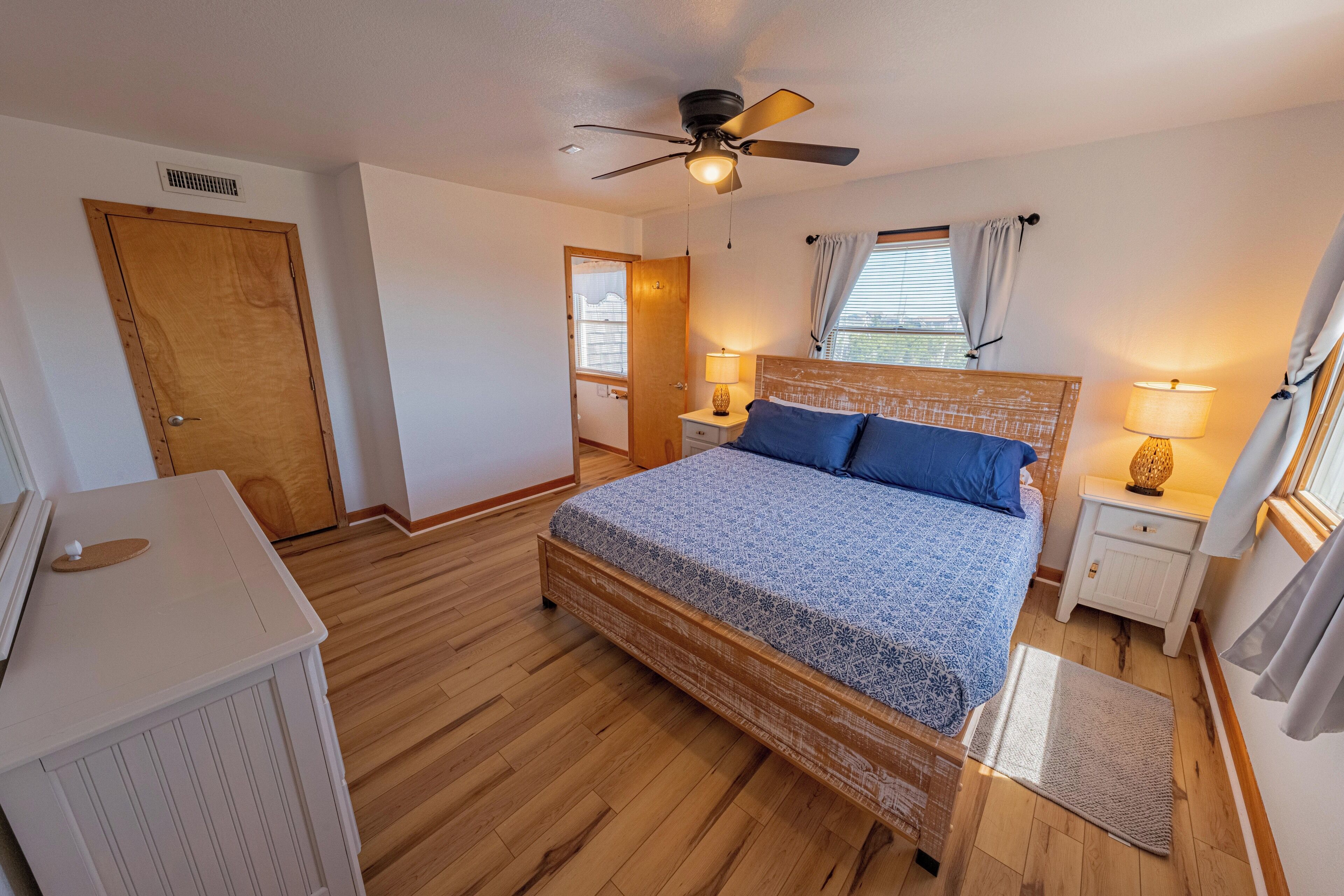 A bedroom with a wooden floor, a bed with a blue and white patterned bedspread, two bedside lamps, a ceiling fan, and a window with curtains.