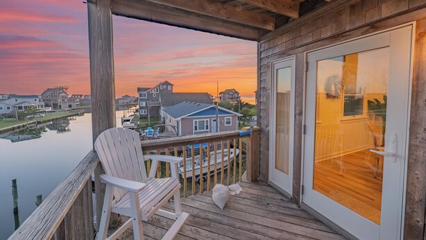 A wooden deck with a white rocking chair, a glass door, and a view of houses and water. - Spacious, newly remodeled, canal front home located in Avon, NC. (Avon)