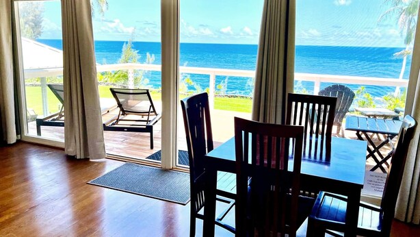 A room with a large sliding glass door leading to a balcony with a view of the ocean. There are two chairs and a small table on the balcony. - The Treehouse (Hakalau)