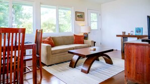 A living room with a sofa, a coffee table, and a TV stand. There are windows with a view of greenery outside. - The Treehouse (Hakalau)