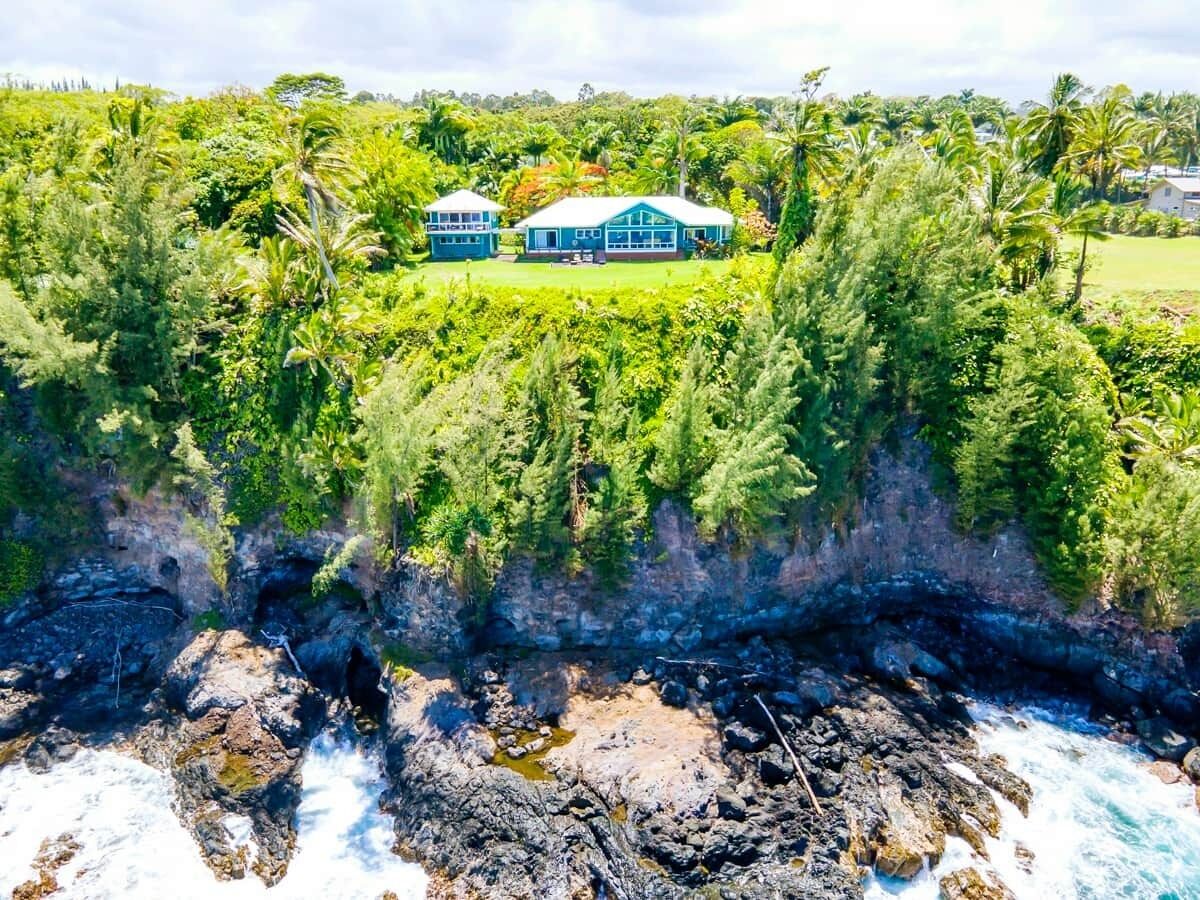 A coastal property with two blue buildings surrounded by lush greenery and a rocky shoreline.