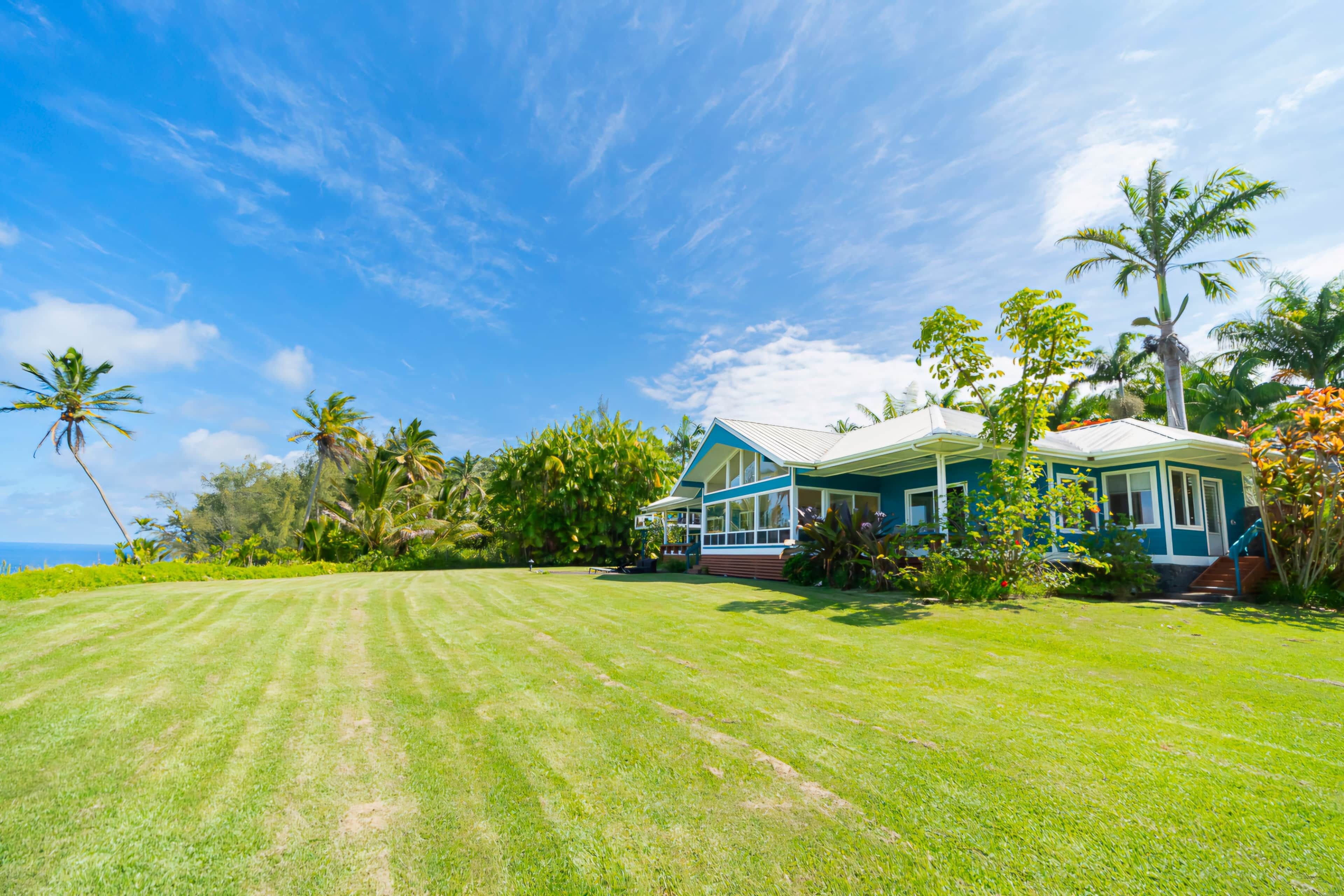 A large, two-story house with a blue exterior and white trim, surrounded by a well-maintained lawn and tropical vegetation.