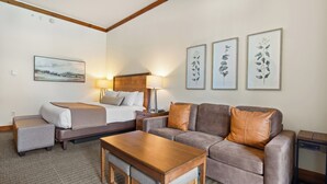 A hotel room with a bed, a sofa, a small table, and a lamp. There are framed botanical prints on the wall. - Studio Spruce Peak Ski In Ski Out "La Voûte" #147 (Stowe)