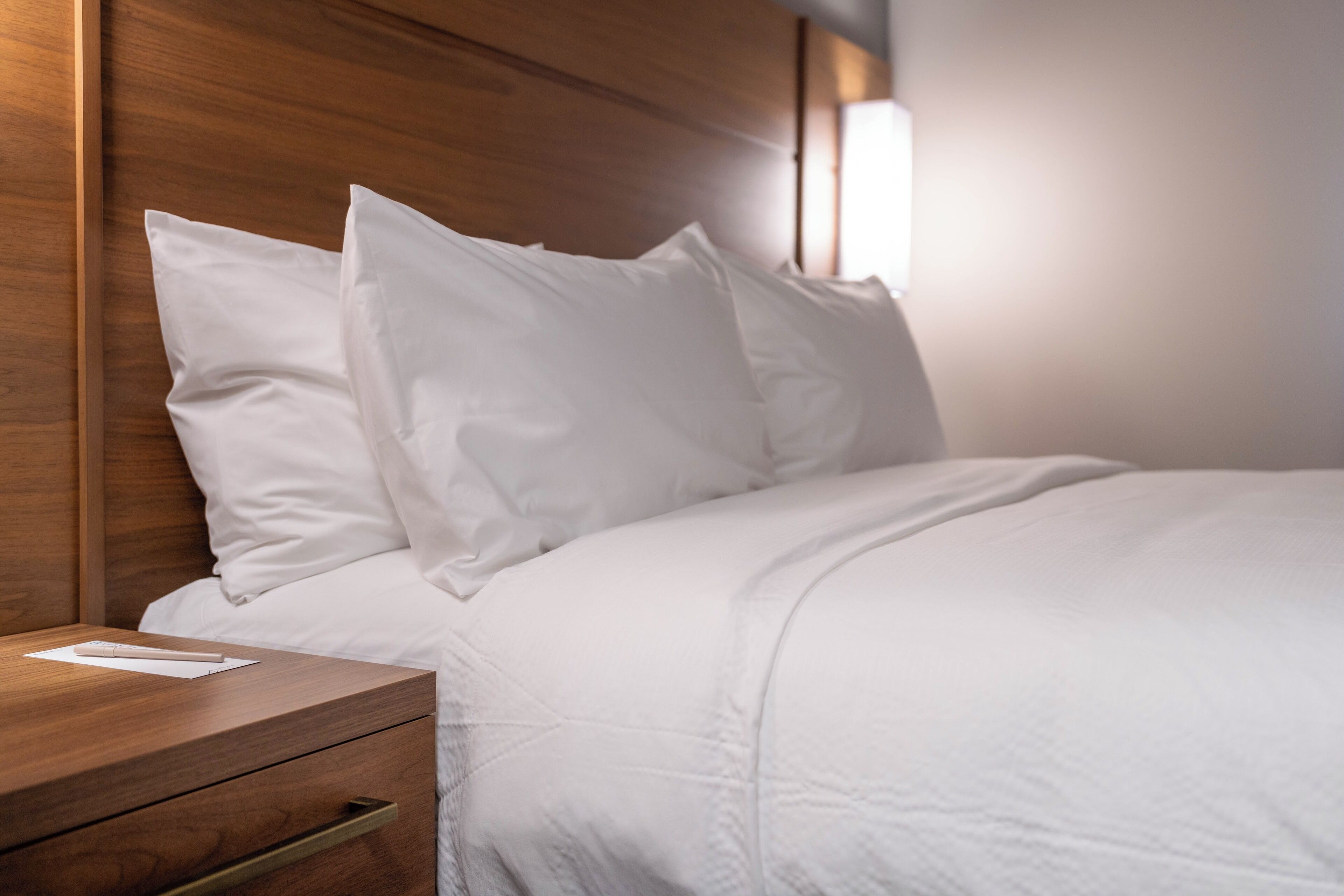 Un lit bien fait, avec du linge de lit blanc et des oreillers, une table de chevet en bois dotée d’un tiroir et un luminaire fixé au mur.