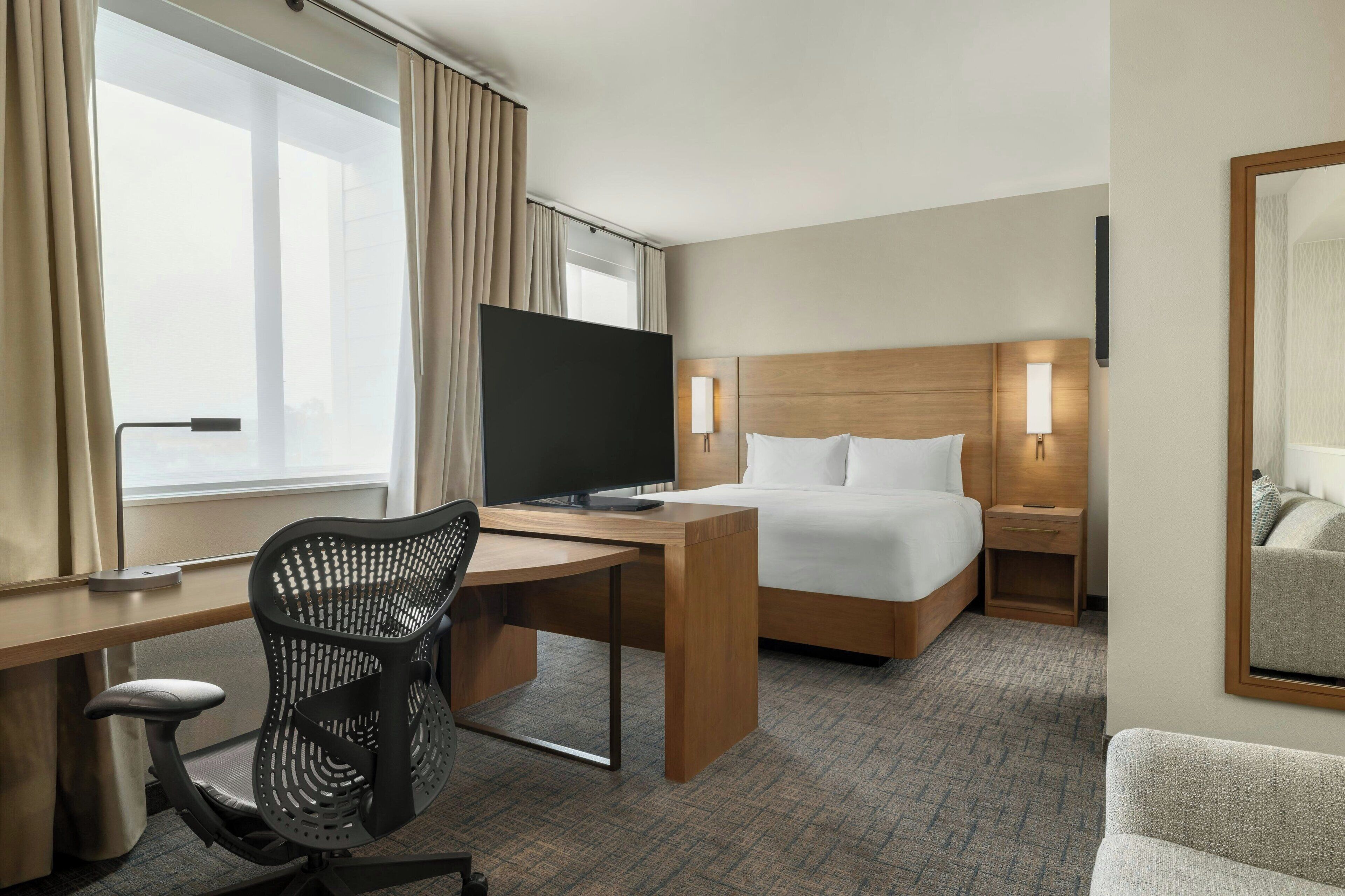 A hotel room with a bed, a desk with a computer, a chair, a TV, and a window with curtains.