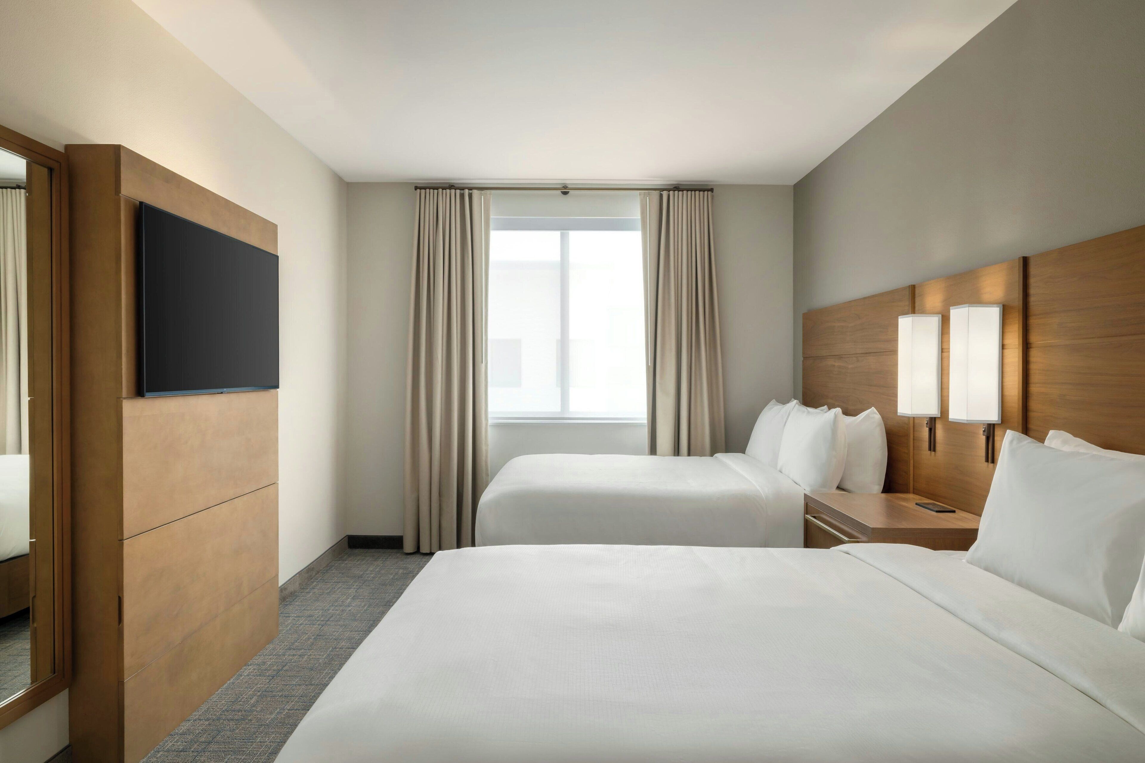 Une chambre d’hôtel avec deux lits, un téléviseur à écran plat, une tête de lit en bois et une fenêtre avec des rideaux.