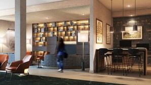 A modern lobby with a central seating area, a long bookshelf, and a dining setup with multiple chairs and tables.