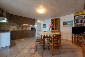 Una cucina con mobili in legno, un forno a microonde e un tavolo da pranzo con sedie. Si nota una libreria con libri e un quadro appeso al muro.