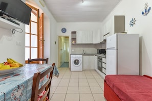 A compact kitchen with a washing machine, a dining table with fruits, and a TV mounted on the wall.