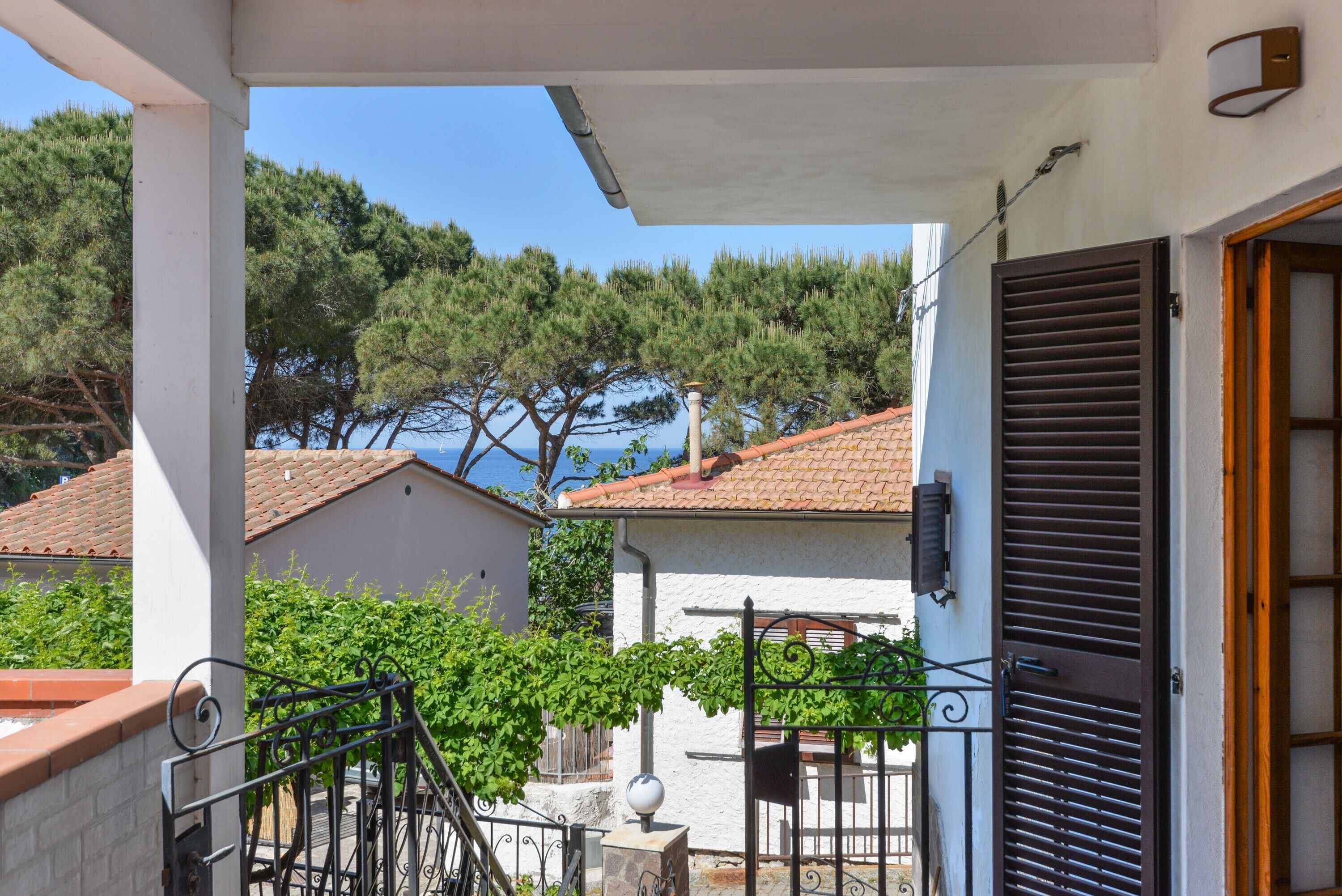 A balcony with a view of a building and trees, a metal gate, and a door with shutters.