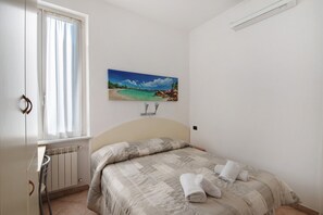 Un dormitorio con cama, ventana con cortinas, radiador, lámpara de pared y un cuadro de una playa.