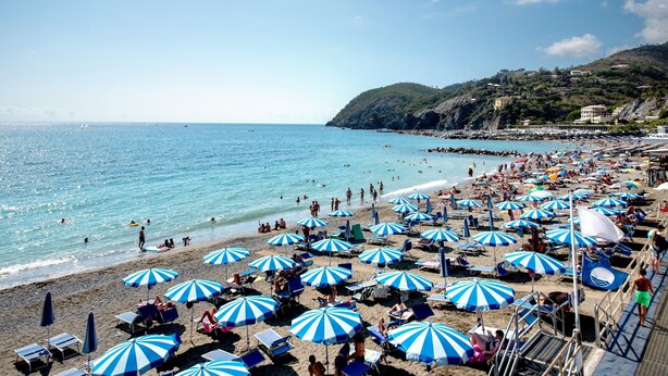 Une plage parsemée de nombreux parasols rayés de bleu et de blanc, une étendue de sable et une côte rocheuse.