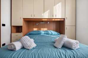 A neatly made bed with blue bedding and two rolled towels, a wooden headboard, and a small bedside table.