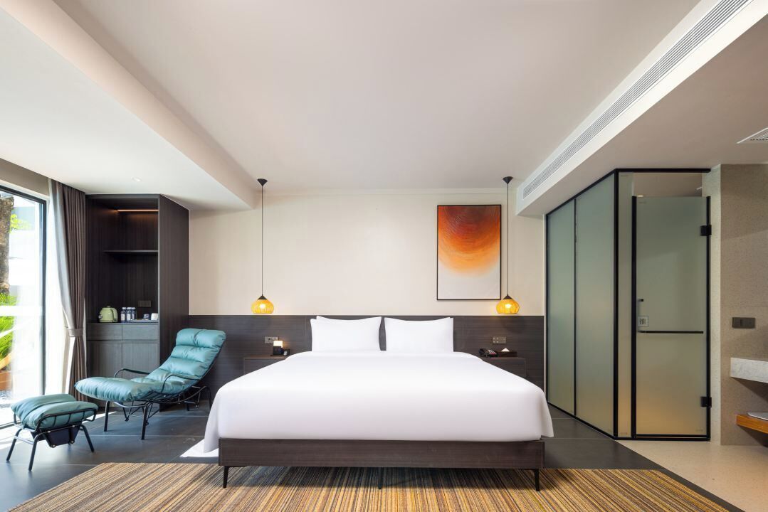 Une chambre d’hôtel moderne dotée d’un grand lit, d’un coin salon avec un fauteuil, d’une armoire et d’une grande fenêtre offrant une vue.