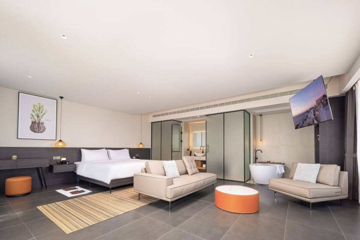 Quarto de hotel moderno com uma cama, dois sofás, um banquinho redondo laranja, banheiro com banheira e um espelho grande.