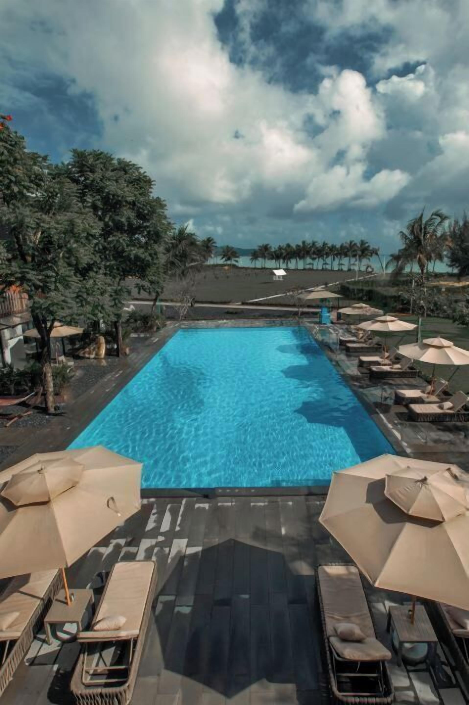 Ein rechteckiger Pool mit klarem, blauem Wasser, umgeben von Liegestühlen und Sonnenschirmen, im Hintergrund Palmen.