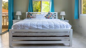 Una cama blanca con cabecera de madera, dos mesitas de noche con lámparas y una ventana con cortinas azules.