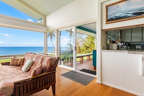 A room with a wooden floor, a bed with a floral bedspread, a view of the ocean, a kitchen with a countertop, and a sliding glass door leading to an outdoor area with palm trees. - The Cliffs (Hakalau)