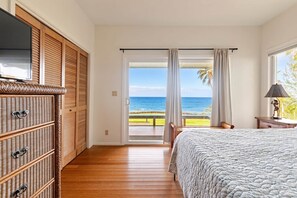Ein Schlafzimmer mit einem Bett, einem Fernseher, einer Holzkommode und Blick auf den Ozean durch eine Schiebetür.