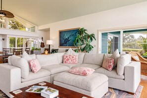 A living room with a sectional sofa, a coffee table, a dining area with chairs, a guitar, and a large window with a view of greenery. - The Cliffs (Hakalau)