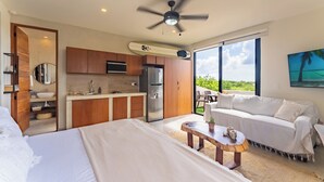 A modern bedroom with a large bed, a small table, and a sofa. There is a kitchen area with a refrigerator and a microwave. The room has large windows with a view of the outdoors. - Studio 55 Private HotTube (Puerto Aventuras)