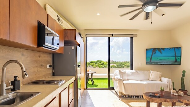 A modern kitchen with wooden cabinets, a built-in microwave, and a large window leading to a balcony with a view of greenery. - Studio 55 Private HotTube (Puerto Aventuras)