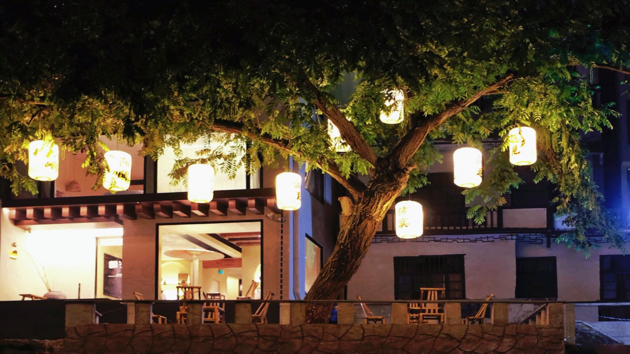 A tree with hanging lanterns in front of a building with a stone wall and outdoor seating.