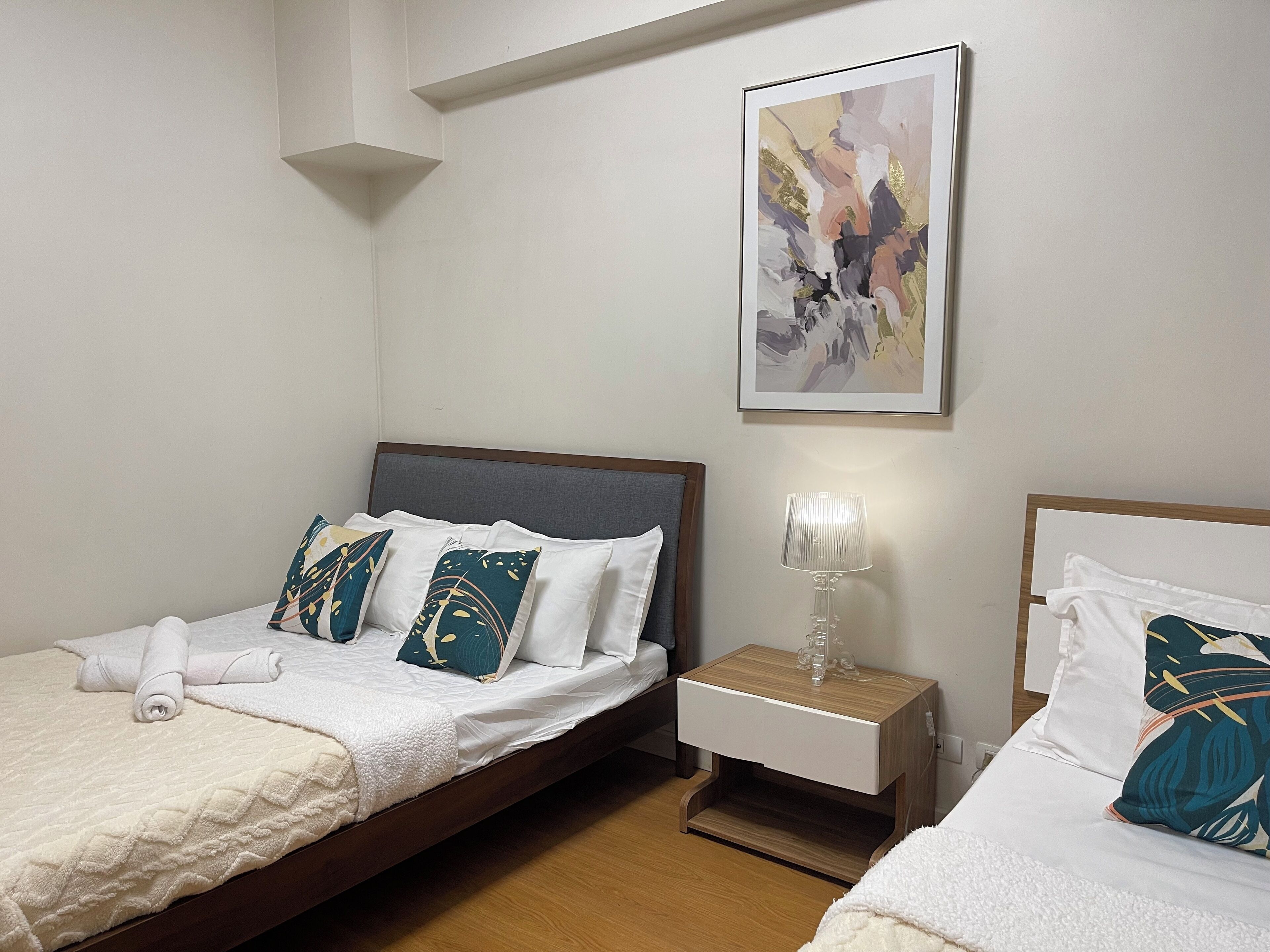 Ein Hotelzimmer mit zwei Betten, einem Nachttisch mit Lampe und einem Bilderrahmen mit einem abstrakten Gemälde an der Wand.